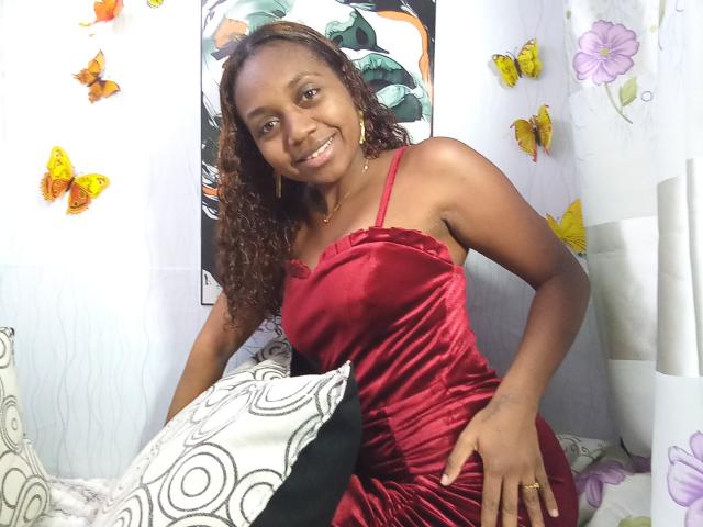 LindahSexy, a dark-skinned woman with curly brown hair, wearing a red velvet dress, smiling at the camera, sitting on a bed with colorful butterfly decorations.