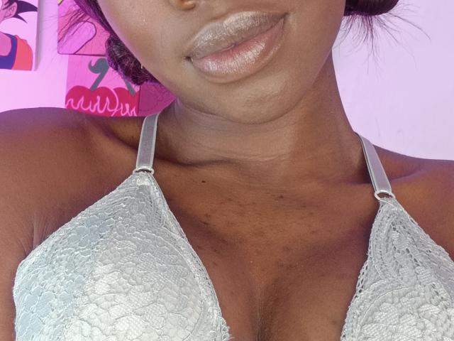 Close-up of Nahomyyy's chest and lower face. Nahomyyy has dark brown skin and shiny, glittery pink lipstick. She wears a light gray lace bra with silver straps. The bra's lace texture is detailed and patterned. Her collarbone and upper chest are visible. Her hair is slightly curly and dark. The background is pink with a small, colorful illustration on the left. The image focuses on her upper body and lips.