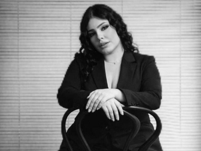 NathalyRose69 with curly dark hair, wearing a black blazer and deep cleavage, sitting with hands on chair, looking slightly to the side, black-and-white photo.