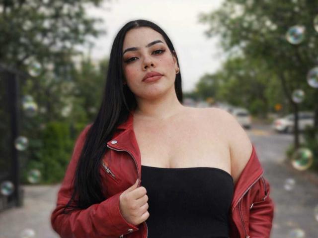 NathalyRose69 with long black hair, red leather jacket, and black strapless top, holding jacket, showing her big ass and small tits, surrounded by floating bubbles.
