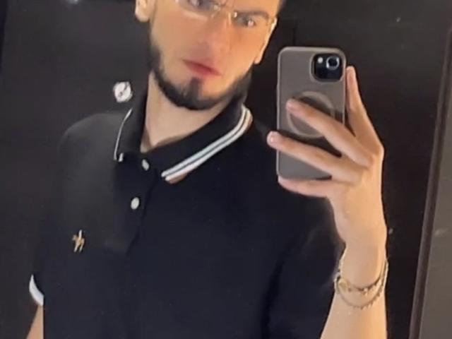 NastyX6 takes a selfie with a smartphone in a dark room. He has a light skin tone, short dark hair, and a beard. Wearing clear glasses, he's dressed in a black polo shirt with white trim and a small emblem on the left chest. His right hand holds the phone, which has a circular ring on the back. A gold bracelet is on his right wrist. The image is slightly blurry.