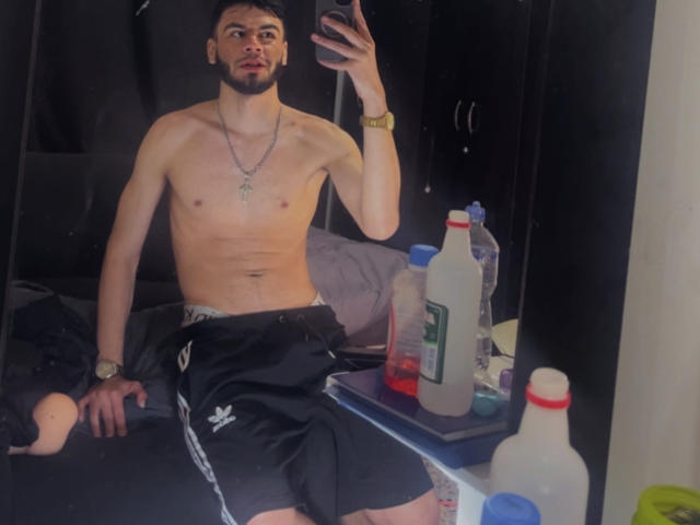 NastyX69, shirtless, holding phone in mirror selfie, black shorts, necklace, watch, bottle on bed, black wardrobe, bottles on table, short dark hair, beard, serious expression.