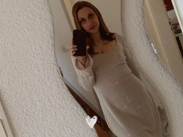 Joie-20-hot with medium tits in a white, ribbed dress, holding a black phone, taking a mirror selfie, with a heart sticker near her ass.