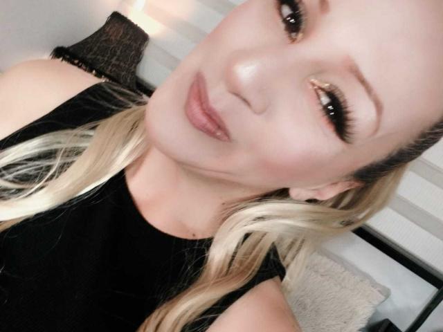 SammyDesire has blonde hair, long eyelashes, and is wearing a black top. She's smiling slightly with glossy pink lips and a subtle makeup look. Her ass is not visible.