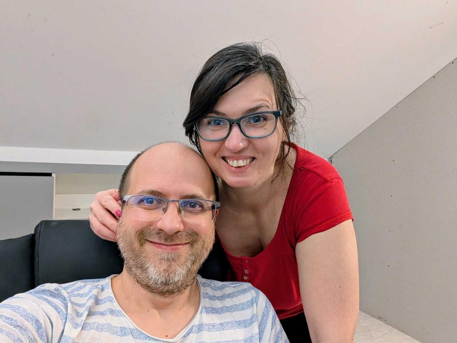 VikAndKarina com óculos, camisa vermelha e seios, apoiada em VikAndKarina, careca com camisa listrada e nádegas. Ambos sorrindo, a mão de VikAndKarina na cabeça de VikAndKarina..