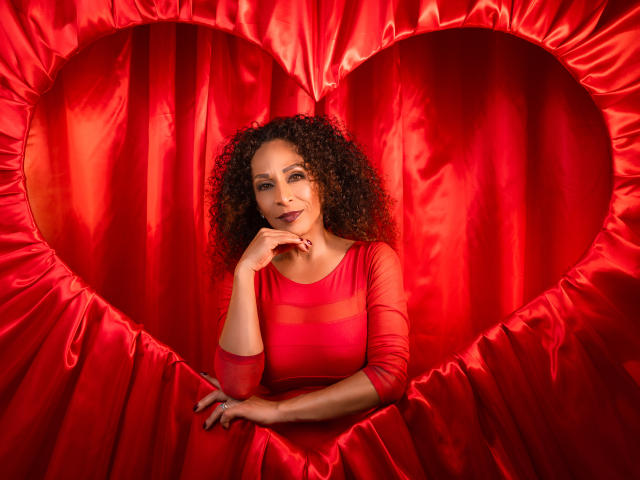 KhazandraX with curly brown hair, red dress, and finger on chin, standing in front of a red heart-shaped satin backdrop, smiling subtly. Her tits are modest, her pussy is unseen.