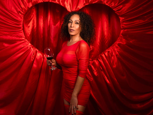 KhazandraX with curly brown hair in a tight red dress holding a glass of red wine, standing in front of red satin with a heart-shaped cutout. Boobs, ass.