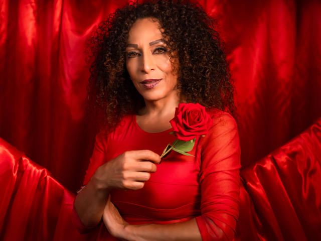 KhazandraX with curly brown hair, wearing a red dress, holding a red rose to her chest, smiling slightly, red satin backdrop, pointing at rose with her right hand.