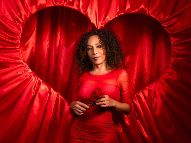 KhazandraX with curly brown hair, wearing a red dress, holding a rose, standing in front of a heart-shaped red satin background, smiling, showing her tits.