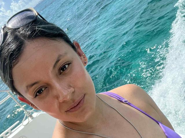 CynthiaMoore, with dark hair in a bun, wearing a purple bikini, has small tits, a silver necklace, and sunglasses on her head, looking at the camera, on a boat.