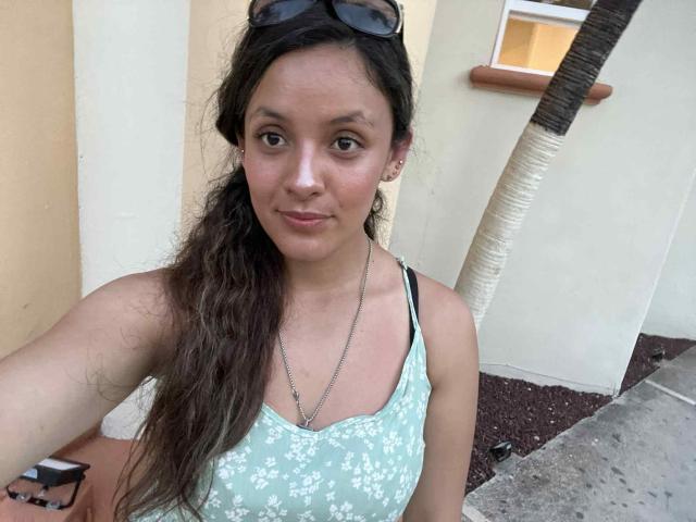 CynthiaMoore with wavy brown hair, wearing a green floral tank top, black bra straps, silver necklace, and sunglasses on her head, standing in front of a yellow wall.