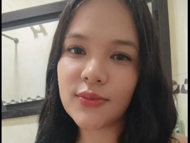 Close-up of AlahiaSexy with medium skin tone and black, wavy hair. She has small, slightly upturned nose, full lips with pink lipstick, and dark, almond-shaped eyes. Her expression is neutral with a slight smile. She is looking directly at the camera. The background shows a bathroom with a mirror and shower area. The lighting is soft, highlighting her skin's natural texture.