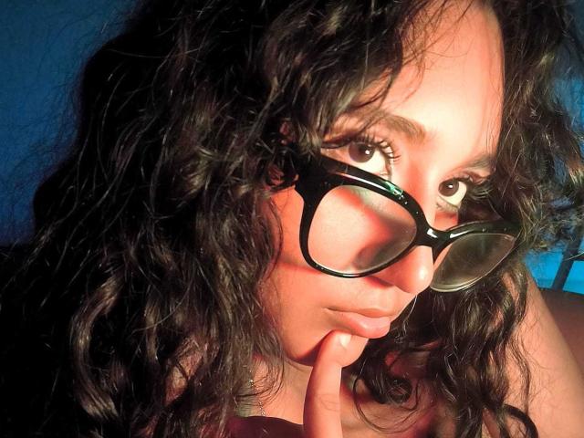 Close-up of AmelieRose with curly black hair, wearing black-rimmed glasses. She has light brown skin and dark eyes. Her right index finger is lightly touching her chin. The light casts shadows on her face, highlighting her cheekbones and the curve of her nose. The background is a dark blue. The image is sharply focused on her facial features and glasses. Natural light.