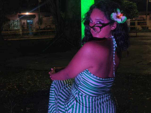 AmelieRose stands in a dark outdoor setting, illuminated by a green light. She has curly black hair, a white and pink flower in her hair, and wears black glasses. She looks over her shoulder, wearing a green and white striped halter dress. The green light casts a purple glow on her skin. She holds her dress with her right hand. The background is mostly dark, with a large tree and a building in the distance. The image has a candid, nighttime feel.