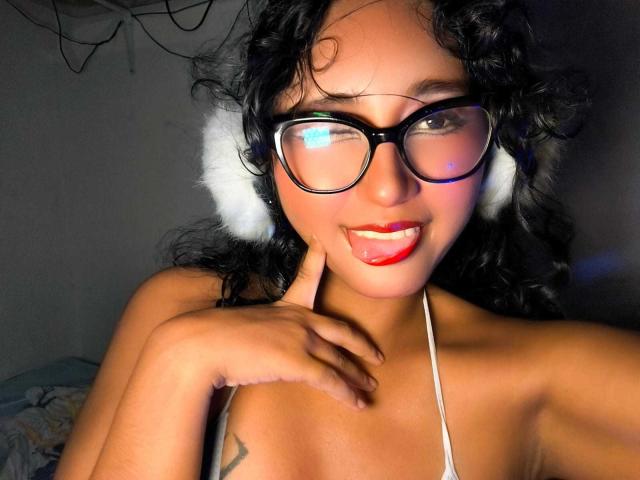 AmelieeRose with curly black hair, glasses, white ear fluffs, red lipstick, tongue out, finger on chin, tan skin, small tattoo on chest, strap visible, smiling.