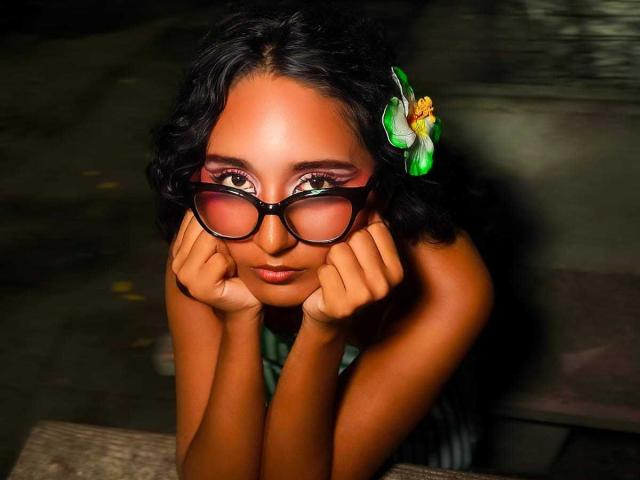 AmelieRose with dark curly hair, green flower, and black glasses, resting her chin on her hands, looking up with pink eyeshadow and brown eyes, in a dimly lit setting.