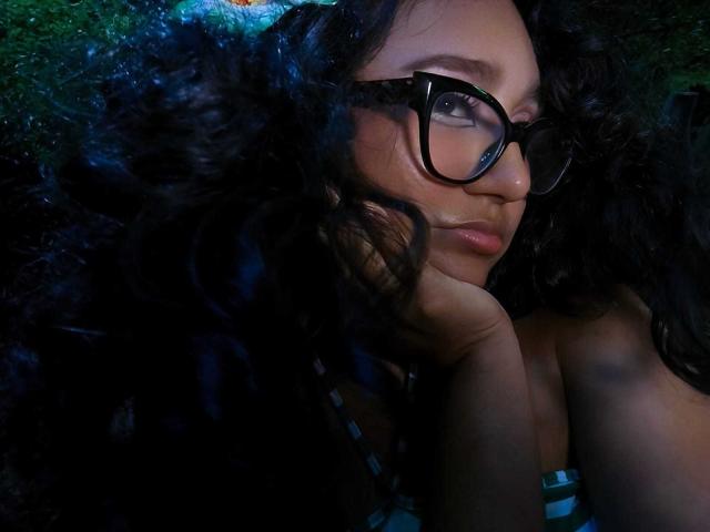 AmelieRose with dark curly hair fills the frame, looking over her shoulder. She wears black, thick-rimmed glasses and a striped top. Her right hand rests under her chin. The lighting is dark, with a greenish-blue glow highlighting her hair. Her expression is neutral, lips slightly parted. The texture of her curly hair contrasts with the smooth skin of her face. Her eyes are focused, giving a sense of depth. The image is close-up, focusing on her face and upper torso.