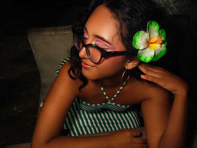 AmelieRose with dark curly hair, green-striped dress, and black glasses, has a green and white flower in her hair, smiling, with her hand near her ear.