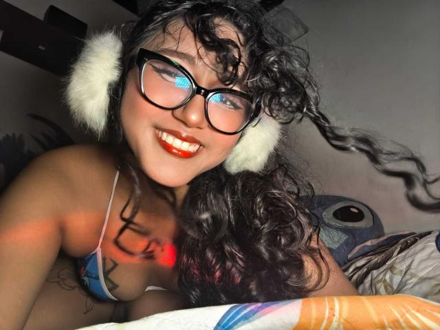 AmelieRose with curly black hair, wearing white furry headphones and black glasses, smiling with red lipstick, blue bikini top, and tattoo on chest, blue eyes glowing.