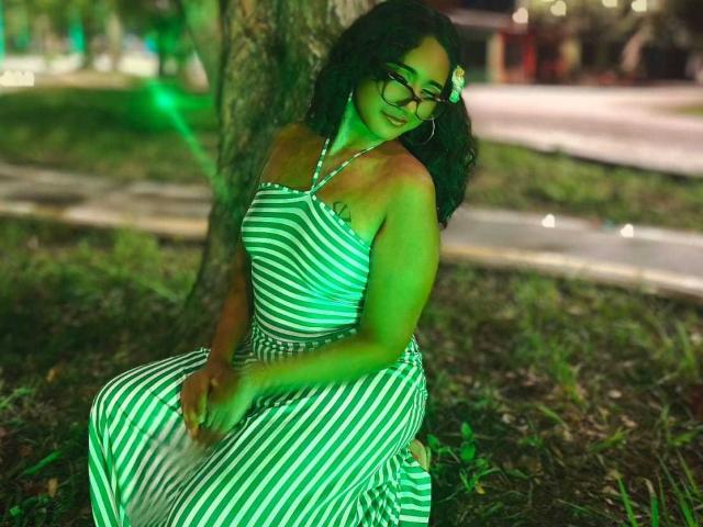 AmelieRose with green-striped dress, sitting against a tree, green light, curly dark hair, flower in hair, glasses, tattoo on shoulder, hands on lap, grassy ground.