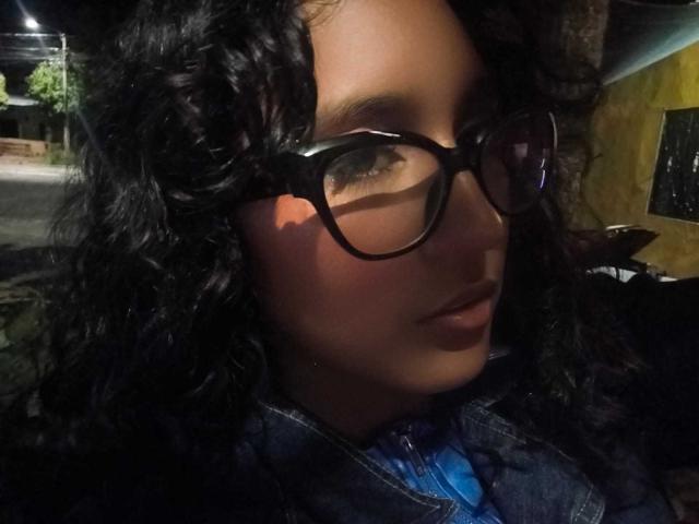 Close-up photo of AmelieeRose with curly black hair, wearing dark glasses with thick frames, and a blue shirt under a dark jacket. Her brown eyes are slightly squinted, and her lips are slightly parted. The light is low, creating shadows on her face. The texture of her curly hair is prominent, and the glasses reflect some light. The background is dark and indistinct. The image has a candid, nighttime feel.