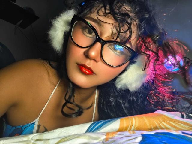 AmelieeRose has curly black hair, wearing large black glasses and white fluffy headphones. Her skin is light brown, with bright red lipstick. She's looking at the camera with a slight smile. She's wearing a blue and white patterned top. The image has a colorful light reflection on her right side. The bed she's on has a colorful patterned blanket. The photo has a close-up, slightly angled perspective.