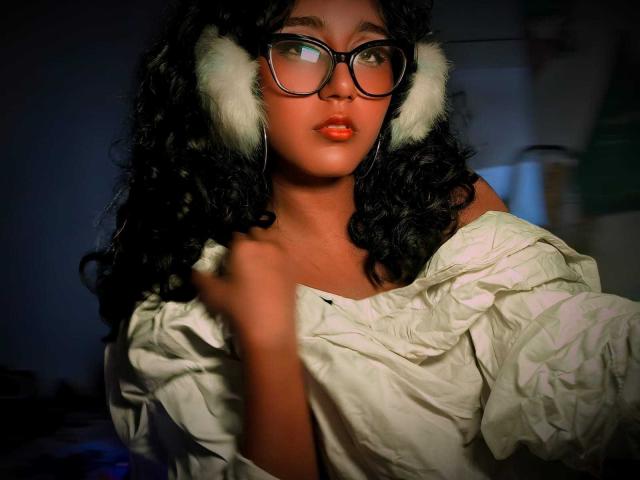 AmelieRose has curly black hair and wears large, black-framed glasses. She's dressed in a loose, white, off-shoulder shirt. Her right hand touches the shirt while her left hand is out of the frame. She has full red lips and a slightly serious expression. White, furry earmuffs cover her ears. The light source is from the right, creating shadows on her left side. The image has a warm, slightly dark tone. The composition centers AmelieRose's face, highlighting her glasses and lips.