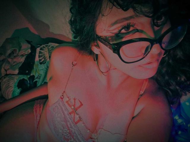 AmelieeRose with curly dark hair, black glasses, pink lace bra, showing her tits, hoop earrings, and a slight pout, under red and green lighting.