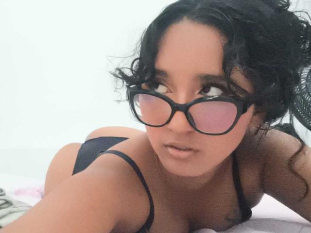 AmelieeRose with curly black hair, wearing black glasses and black bra, lying on bed, looking up, showing her tits and ass, slightly parted lips, soft expression.
