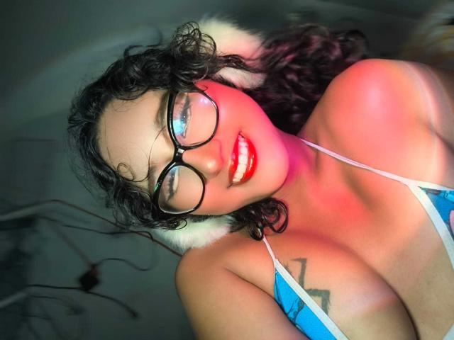 AmelieRose with curly black hair, glasses, red lipstick, and blue top showing her big tits, a tattoo on her right chest, and a white fur trim. Bright red light.