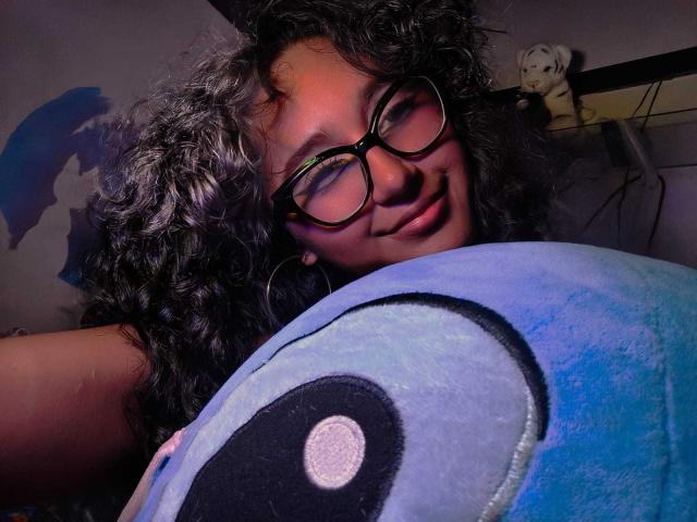 Close-up photo of AmelieeRose with curly black hair and large black glasses. She's smiling slightly, looking into the camera. She's holding a large, blue, plush toy with a black eye and white circle. Her skin is medium brown. The toy covers her chest. A white stuffed animal is in the background on a shelf. The lighting is warm, casting shadows on the wall. AmelieeRose's expression is relaxed and playful. The image has a casual, intimate feel.