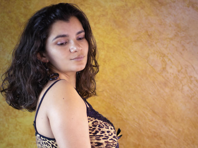 BulmaHot with wavy dark hair, light skin, and a subtle smile, wearing a leopard print top, showing her tits. She has pink eyeshadow and small earrings.