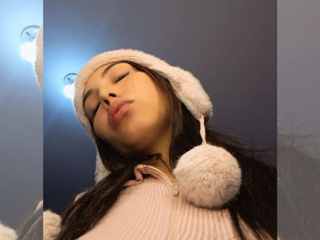 NailaIsaa has her eyes closed, wearing a fluffy white hat with a pom-pom, and a pink ribbed top, her lips are slightly pouted. She's looking down at the camera.