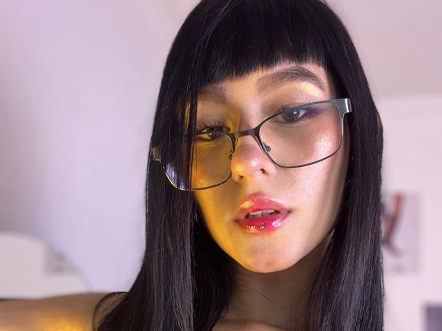 Lycoris with black straight hair, glasses, shiny pink lips, and visible nose piercing, looking slightly to the side, has a soft, glowing skin tone.