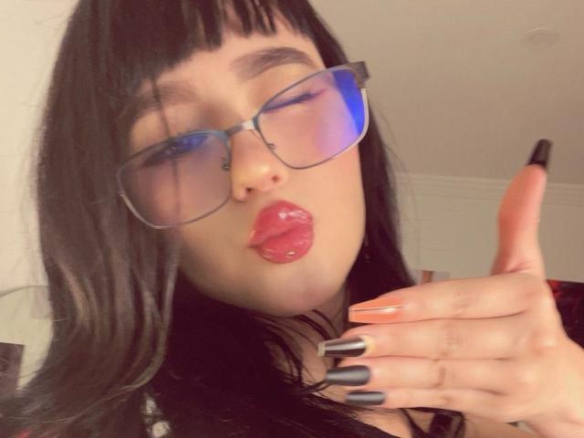 Lycoris with black hair, glasses, red lipstick, and gray nails, giving a thumbs-up, showing a pierced lip, and blue light reflection on her face.