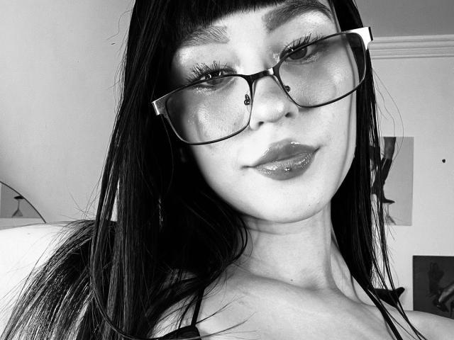 Black-and-white selfie of Lycoris with shiny lips, dark straight hair, and thick glasses, showing her shiny face and subtle smile. Her tits are partially visible.