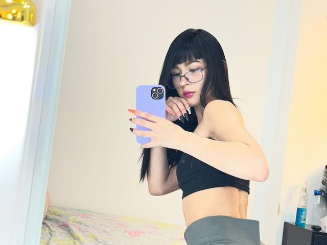Lycoris with long black hair, glasses, and pink lipstick, taking a mirror selfie with a purple phone, showing her black sports bra and grey pants, exposing her flat ass.