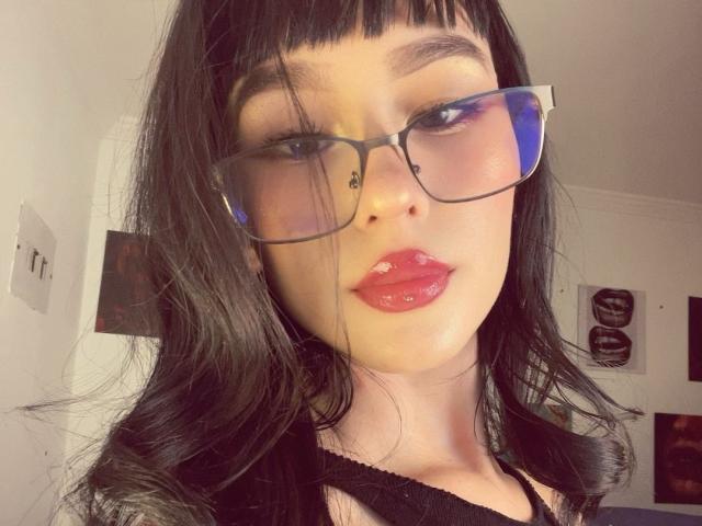 Lycoris has dark, wavy hair, glasses with blue reflections, and shiny pink lips. Her face is slightly angled with a subtle, confident smile. She's wearing a black top.