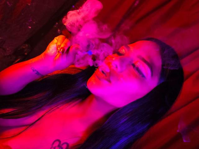 MiaBellucci with long black hair, eyes closed, smoking with pink and purple neon lights. Her tits are partially visible, and she has a tattoo on her left arm.