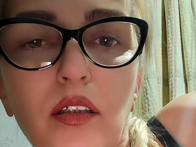 SexyCoco's face, wearing black glasses, with red lipstick, slightly open mouth showing teeth, blonde hair, and fair skin, in a close-up selfie. Boobs partially visible in black top.