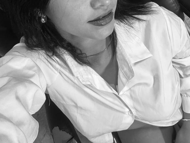 CaritoHam with dark lipstick, wearing a white button-up shirt, choker, and pearl earrings, shot from above. Boobs partially visible, shirt slightly open, showing cleavage. Black-and-white photo.
