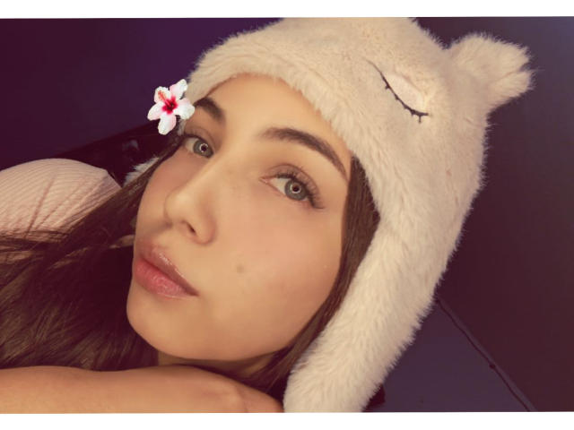 NailaIsaa's face with a cute white fluffy hat featuring a cat face, a pink and white flower sticker on her left cheek, and her brown eyes looking at the camera.