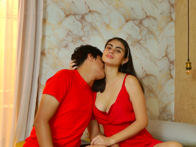 SofiNdLogan in a red dress with cleavage, sitting, her dark-haired boyfriend kissing her neck, wearing a red shirt, in a cozy room with marble-patterned wallpaper.