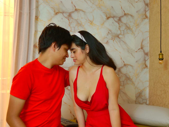 SofiNdLogan with black hair in a red dress, deep cleavage, leaning towards a man in a red shirt, both gazing at each other, in a warmly lit room.