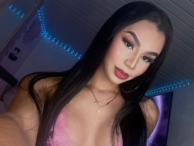 OriannaSimons has long, straight black hair, wearing a pink lace bra that reveals a modest cleavage. She has thick, dark eyebrows, long eyelashes, and red lipstick. She wears gold hoop earrings and a gold necklace with a small pendant. Her skin is light, and she has a confident expression. The image focuses on her upper body and face, with blue LED lights in the background. The ceiling is white and has a grid pattern. The lighting is even, highlighting her facial features and makeup.