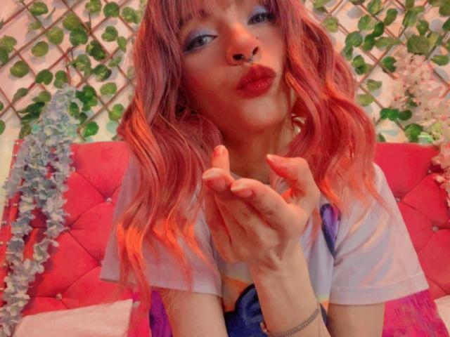 LaurenEvie69 with pink hair, pouty red lips, blue eyeshadow, hands near face, wearing a purple and blue top, looking cute, sitting on a pink tufted bed.