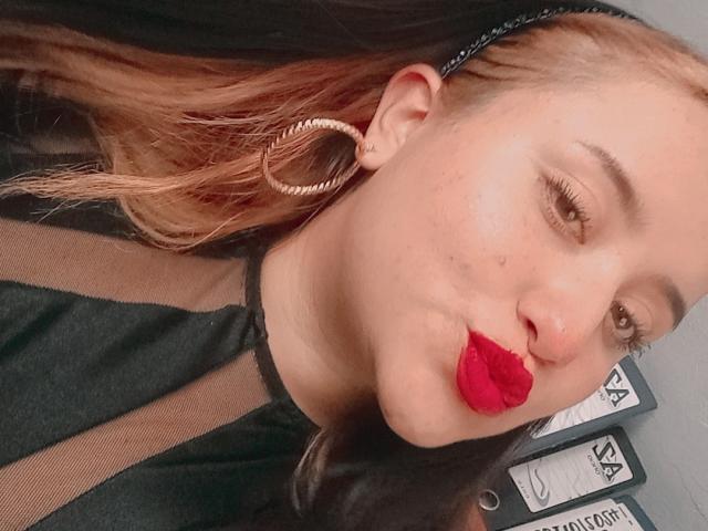 GabrielaMora con rossetto rosso, che bacia l'obiettivo, indossa una maglia a rete nera, orecchini a cerchio dorati, capelli biondi con punte marroni, leggere lentiggini, pelle chiara, trucco leggero, selfie casual..