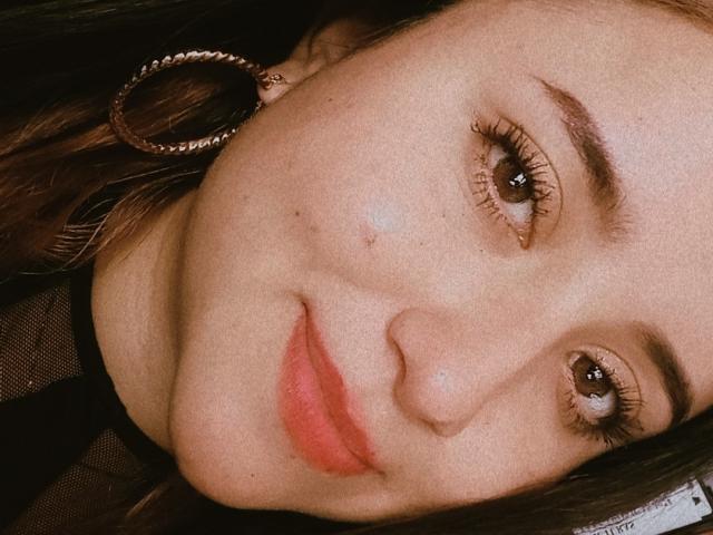 Das Gesicht von Gabriela Mora mit braunen Augen, roten Lippen und einer kleinen Pickel auf der Nase. Sie trägt einen metallenen Ohrring, und ihr braunes Haar ist teilweise sichtbar..