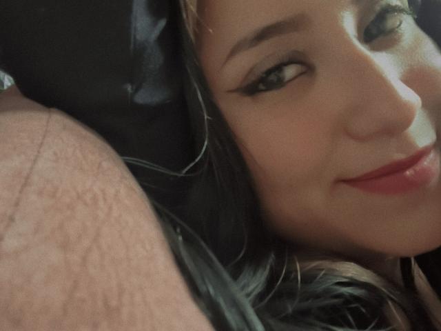 Gabriela Mora, com delineador preto, batom vermelho e cabelo castanho, sorri suavemente ao lado do ombro peludo de um homem. Seu rosto está próximo, mostrando sua bochecha direita..