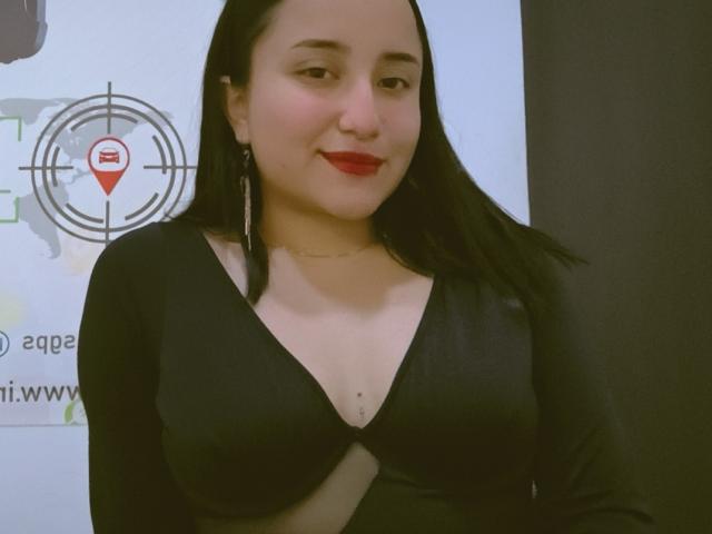 Gabriela Mora, con capelli neri, rossetto rosso, vestito nero a V che mette in mostra il seno, di media taglia, indossa orecchini pendenti argentati, sorride, ha la pelle chiara, una corporatura media, ed è in piedi di fronte a un muro..